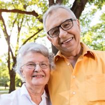 Ein älteres Paar lächelt im Freien mit Bäumen im Hintergrund und verkörpert Glück und Ruhe. Der Mann, der eine Brille und ein orangefarbenes Hemd trägt, und die Frau mit kurzen grauen Haaren und einer weißen Bluse scheinen diese friedlichen Momente gemeinsam zu genießen, während sie über Dinge wie Erbrecht diskutieren.