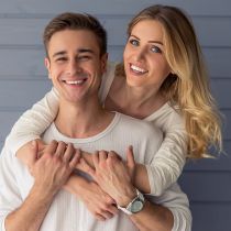 Ein lächelndes Paar posiert vor einer blauen Holzwand. Die Frau hat blonde Haare und umarmt den Mann von hinten. Beide tragen legere, helle Kleidung. Der Mann hält mit fröhlichem Gesichtsausdruck ihre Arme und zeigt eine Armbanduhr an seinem linken Handgelenk.