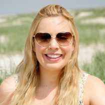 Eine lächelnde Frau mit langen blonden Haaren und einer herzförmigen Sonnenbrille steht im Freien vor Sanddünen und grünem Gras und genießt den Sonnenschein, nachdem sie ihre Handelsregisteranmeldung bei einem Notar in Berlin abgeschlossen hat, mit einem klaren Himmel im Hintergrund.