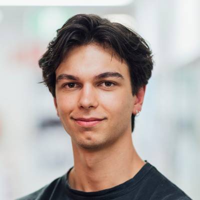 Ein junger Mann mit kurzem, dunklem Haar und einem Ohrring lächelt leicht in die Kamera. Er trägt ein schwarzes Hemd, und der leicht verschwommene Hintergrund suggeriert einen hellen Innenraum - ideal für eine Online-Notarsitzung.