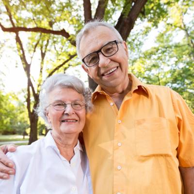 Ein älteres Paar steht im Freien dicht beieinander und lächelt in die Kamera. Der Mann trägt eine Brille und ein orangefarbenes Hemd, während die Frau in einer weißen Bluse ihr Telefon in der Hand hält - vielleicht feiern sie ihre erste Erfahrung mit einem Online-Notar. Bäume und Sonnenlicht erhellen die Szene.
