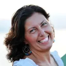 Eine Frau mit gewelltem braunem Haar lächelt breit in die Kamera. Sie trägt Ohrringe und ein weißes Oberteil. Sie steht vor etwas, das aussieht wie die verschwommene Fassade eines Grundbuchamtes an einem sonnigen Tag.