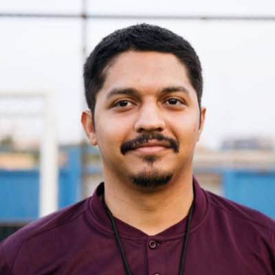 Ein Mann mit kurzen dunklen Haaren, Gesichtsbehaarung und Schnurrbart, der ein kastanienbraunes Hemd und ein schwarzes Schlüsselband trägt, steht im Freien vor einem unscharfen blauen und weißen Hintergrund und scheint bereit zu sein, als Online-Notar zu helfen.