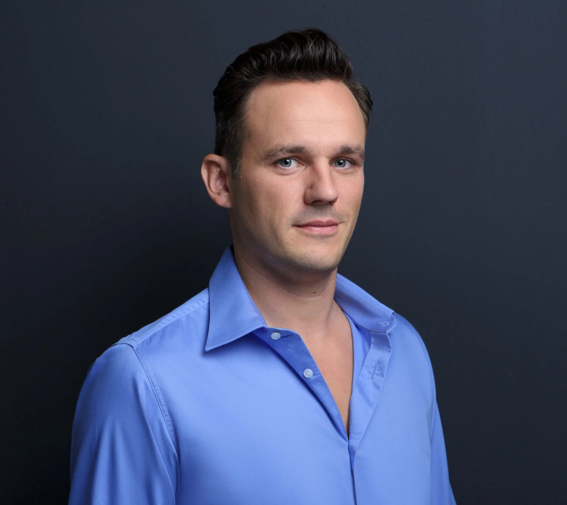 Grischa Sebastian Franke, Rechtsanwalt und Notar in Berlin, ein Mann mit kurzen dunklen Haaren, der ein hellblaues Hemd trägt, steht vor einem schlichten dunklen Hintergrund und blickt mit neutralem Blick leicht zur Seite während eines Online Notar Termins zu einem Geschäftsführerwechsel.