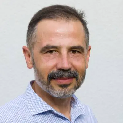 Ein Mann mittleren Alters mit kurzen braunen Haaren, einem grauen Bart und Schnurrbart lächelt leicht. Er trägt ein hellblau gemustertes Hemd und posiert vor einem hellen Hintergrund - vielleicht bereit, über Themen wie die Gründung der Stammkapital UG zu sprechen.