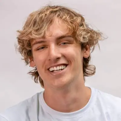 Eine junge Person mit gewelltem blondem Haar und hellem Teint lächelt in die Kamera. Sie trägt ein schlichtes helles Hemd, als wäre sie bereit, einen Gesellschaftsvertrag oder ein Musterprotokoll zu unterschreiben, vor einem neutralen Hintergrund.