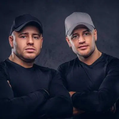 Zwei Männer, die wie Zwillinge aussehen, stehen mit verschränkten Armen nebeneinander und tragen passende schwarze Hemden und Baseballkappen - einer schwarz, einer grau - vor einem dunklen Hintergrund und verkörpern den Geist der Gründung von zu Hause.