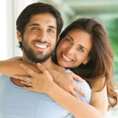 Eine lächelnde Frau mit langen braunen Haaren umarmt einen bärtigen Mann von hinten, beide schauen in die Kamera. Sie wirken glücklich und entspannt in einem hellen Innenraum, vielleicht feiern sie, nachdem sie erfolgreich Firmennamen gewählt haben.