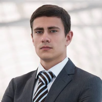 Ein junger Mann in einem Geschäftsanzug mit gestreifter Krawatte steht selbstbewusst mit verschränkten Armen da. Er hat kurzes dunkles Haar und einen ernsten Gesichtsausdruck. Der Hintergrund ist unscharf und hell und suggeriert eine moderne Innenraumumgebung.