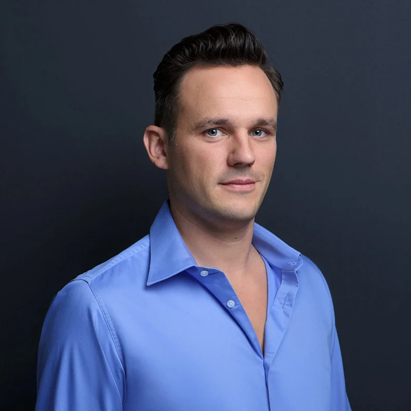 Ein Mann mit kurzen dunklen Haaren trägt ein hellblaues Button-up-Hemd mit offenem Kragen, steht vor einem schlichten dunklen Hintergrund und blickt leicht zur Seite, als würde er über einen Gesellschaftsvertrag oder ein Musterprotokoll nachdenken.