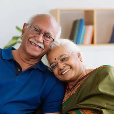 Ein älteres Ehepaar sitzt in einem Haus zusammen und lächelt sich herzlich zu. Die in einen grünen Saree gekleidete Frau stützt ihren Kopf auf die Schulter des Mannes. Im sanft verschwommenen Hintergrund sind Bücherregale zu sehen, die an Geschichten der ausländischen Gesellschafter und Geschäftsführer GmbH erinnern.