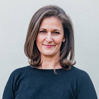 Frau mit schulterlangen braunen Haaren, die ein schwarzes Oberteil trägt und leicht lächelt, während sie vor einem schlichten hellen Hintergrund steht - vielleicht bereitet sie sich auf einen Notartermin vor und überlegt, was sie mitbringen soll.