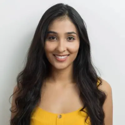 Eine Frau mit langen, gewellten dunklen Haaren lächelt in die Kamera. Bekleidet mit einem gelben ärmellosen Oberteil steht sie selbstbewusst vor einem schlichten weißen Hintergrund, der den Geist von gründung mit digitalem office widerspiegelt.