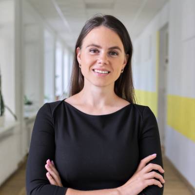 Eine Frau mit langen braunen Haaren, die ein schwarzes Kleid trägt, steht in einem hellen Flur mit verschränkten Armen und lächelt selbstbewusst in die Kamera - eine Verkörperung des Geistes von Holding gründen. Der Korridor hat weiße Wände mit einem gelben Streifen.