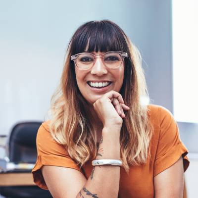 Eine lächelnde Frau mit langen, gewellten Haaren und Brille sitzt in einem Haus und stützt ihr Kinn auf die Hand. Sie trägt ein orangefarbenes Oberteil und hat sichtbare Tattoos auf dem Arm - perfekt für einen Vortrag über die Gründung mit juristischen Personen als Gesellschafter.