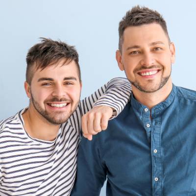 Zwei lächelnde Männer posieren gemeinsam vor einem hellblauen Hintergrund. Der eine, der ein gestreiftes Hemd trägt, stützt seinen Arm auf die Schulter des anderen, der mit einem blauen Button-up bekleidet ist - beide wirken fröhlich und freundlich, möglicherweise diskutieren sie über eine Satzungsänderung GmbH.