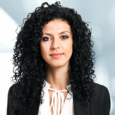 Eine Frau mit langen, lockigen schwarzen Haaren trägt eine weiße Bluse mit Schleife und einen schwarzen Blazer und steht vor einem unscharfen Hintergrund. Sie blickt mit einem leichten Lächeln in die Kamera, bereit, die Ablauf Notartermin GmbH zu erklären.