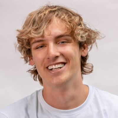 Ein junger Mann mit gewelltem, blondem Haar und hellem Teint lächelt in die Kamera. Er trägt ein weißes T-Shirt vor einem schlichten, hellen Hintergrund und strahlt Selbstbewusstsein aus, wie es sich für eine handdenhaftung GmbH in Gründung gehört.