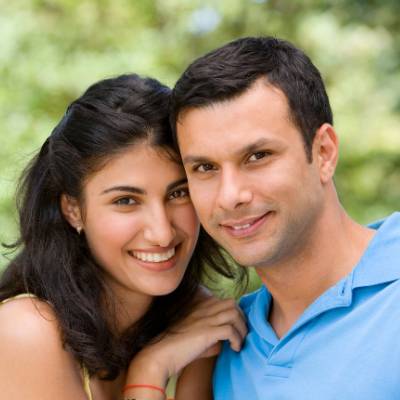 Eine lächelnde Frau mit langen dunklen Haaren lehnt sich liebevoll an die Schulter eines Mannes in einem hellblauen Hemd. Im Freien, vor einem unscharfen, grünen Hintergrund, diskutieren sie über Themen wie Haftung nach der Gründung der GmbH UG.