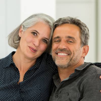 Ein Paar mittleren Alters lächelt und posiert eng beieinander in einem Innenraum, wie mehrere Gesellschafter, die kurz davor sind, eine GmbH zu gründen. Die Frau hat graue Haare und trägt eine marineblaue gepunktete Bluse, der Mann hat kurze graue Haare und trägt ein dunkles Hemd.