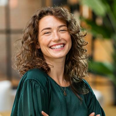 Eine junge Frau mit lockigem braunem Haar und grüner Bluse lächelt strahlend in die Kamera. Sie steht in einem Innenraum mit unscharfen Pflanzen und warmer Beleuchtung im Hintergrund - vielleicht recherchiert sie für die ablehnungsgründe Handelsregister Berlin GmbH.