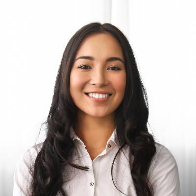 Eine junge Frau mit langen dunklen Haaren, die ein weißes Button-up-Hemd trägt, steht lächelnd vor einem hellen, weißen Hintergrund, der die Klarheit und Offenheit widerspiegelt, die das Transparenzregister fördert.