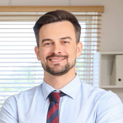 Ein Mann mit kurzen dunklen Haaren und Bart, der ein hellblaues Hemd und eine rot-blau gestreifte Krawatte trägt, lächelt, während er in einem Innenraum vor einem Fenster mit Jalousien steht - vielleicht kurz nach seiner Entscheidung, die Stammkapital einzahlen GmbH UG.