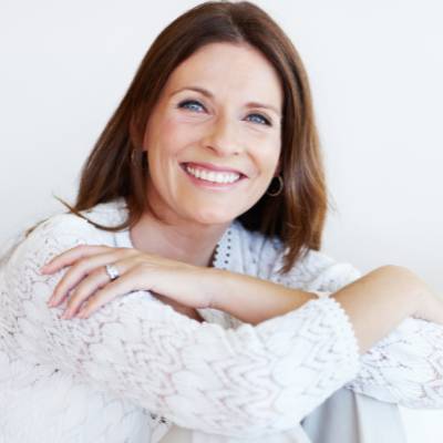 Eine Frau mit langen braunen Haaren, die einen weißen strukturierten Pullover trägt, sitzt mit den Armen auf den Knien und lächelt warm in die Kamera - bereit für ihr nächstes Abenteuer, vielleicht sogar für eine Gewerbeanmeldung in Berlin.