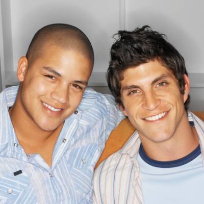 Zwei junge Männer sitzen dicht beieinander und lächeln in die Kamera, entspannt und glücklich in ihren legeren gestreiften Hemden. Der neutrale Innenhintergrund suggeriert einen ruhigen Moment, vielleicht nach ihrer Anmeldung beim Finanzamt nach Gründung.