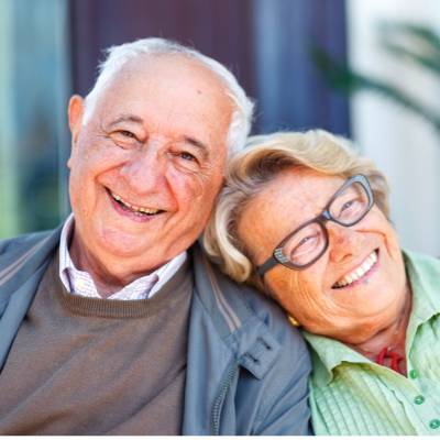 Ein älteres Ehepaar sitzt eng beieinander und lächelt freundlich. Der Mann trägt eine graue Jacke und einen braunen Pullover, und die Frau, die eine Brille und ein grünes Hemd trägt, lehnt ihren Kopf an seine Schulter. Sie wirken glücklich und zufrieden.