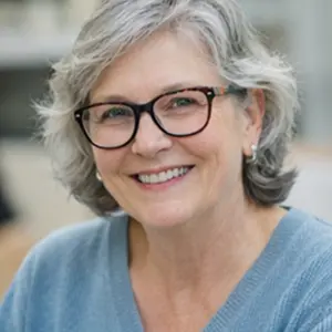 Eine lächelnde ältere Frau mit kurzen grauen Haaren und Brille, die einen hellblauen Pullover trägt, sitzt in einem Innenraum mit unscharfem Hintergrund - vielleicht wartet sie darauf, einen Notartermin der Adressänderung GmbH zu besprechen.