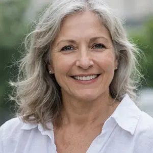 Eine lächelnde ältere Frau mit gewelltem grauem Haar und weißem Hemd steht im Freien vor einem unscharfen grünen Hintergrund.