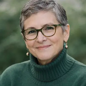 Eine lächelnde Person mit kurzen grauen Haaren, die eine Brille, Perlenohrringe und einen dunkelgrünen Rollkragenpullover trägt, posiert im Freien vor einem verschwommenen grünen Hintergrund - vielleicht in Erwartung eines Notartermins bei der Adressänderung GmbH.