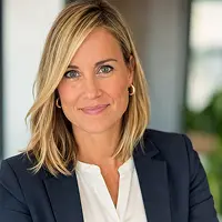 Eine Frau mit blondem Haar, die einen marineblauen Blazer und eine weiße Bluse trägt, lächelt sanft in die Kamera in einer professionellen Innenumgebung, die an ein Büro von Grundschuld Notar Berlin erinnert.