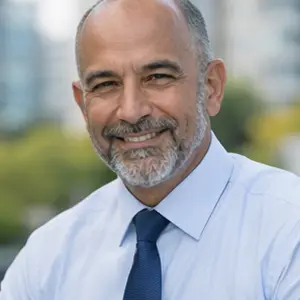 Ein Mann mittleren Alters mit einer Glatze, grauem Bart und Schnurrbart lächelt. Er trägt ein hellblaues Hemd und eine dunkelblaue Krawatte und steht im Freien in der Nähe der Holding gründen Berlin; der unscharfe Hintergrund zeigt grüne Bäume und Gebäude.