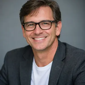 Ein lächelnder Mann mit kurzen braunen Haaren und Brille, der einen grauen Blazer über einem weißen T-Shirt trägt, posiert selbstbewusst vor einem schlichten grauen Hintergrund - bereit für UG gründen.