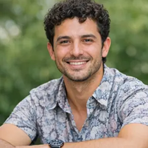 Ein Mann mit lockigem, dunklem Haar und Bart sitzt lächelnd, in einem gemusterten, kurzärmeligen Hemd und mit einer Uhr im Freien in Berlin - vielleicht genießt er eine Pause, nachdem er sich für die GmbH gründen entschieden hat. Der verschwommene grüne Hintergrund trägt zur entspannten Stimmung bei.