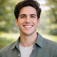 Ein junger Mann mit dunklem Haar lächelt in die Kamera. Er trägt ein grünes Button-up-Hemd über einem weißen T-Shirt. Im Hintergrund sind sanft verschwommene Grünflächen und Sonnenlicht zu sehen, die eine entspannte Berliner Atmosphäre hervorrufen - ideal für alle, die ihren Grundschuld Notar treffen.