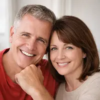 Ein Mann und eine Frau mittleren Alters sitzen eng beieinander in einem Haus, beide lächeln warm in die Kamera - vielleicht feiern sie eine kürzlich erfolgte Grundschuldlöschung. Der Mann hat kurzes graues Haar und trägt ein rotes Hemd, während die Frau braunes Haar hat und ein helles Oberteil trägt.