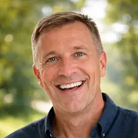 Ein Mann mittleren Alters mit kurzen hellbraunen Haaren lächelt breit und trägt ein dunkelblaues Hemd mit Kragen. Er steht im Freien vor einem unscharfen grünen Hintergrund, vielleicht nach einem Treffen mit einem Notar der Immobilien Schenkung in Berlin.