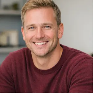 Ein Mann mit kurzen blonden Haaren und gestutztem Bart, der einen kastanienbraunen Pullover trägt, lächelt, während er ein Buch über das grüne Berlin in der Hand hält, das in einem Innenraum mit Regalen und Dekoration im sanft verschwommenen Hintergrund sitzt.