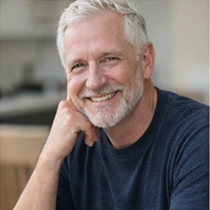 Ein lächelnder älterer Mann mit grauen Haaren und Bart, der ein dunkles T-Shirt trägt, sitzt in einem Haus und stützt seine Hand auf sein Kinn - vielleicht denkt er über die jüngsten Angelegenheiten der Umfirmierung des Notars nach. Der Hintergrund ist sanft verschwommen.