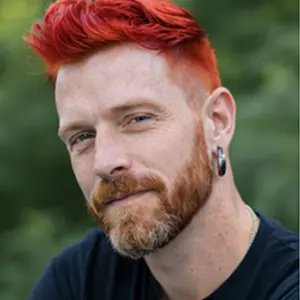 Eine Person mit leuchtend rotem Haar und einem kurzen Bart blickt in die Kamera, trägt ein dunkles Hemd und einen Ohrring. Vor dem verschwommenen Grün verkörpern sie den kreativen Geist Berlins - vielleicht denken sie über ihr nächstes großes Projekt nach oder haben grüne Träume.