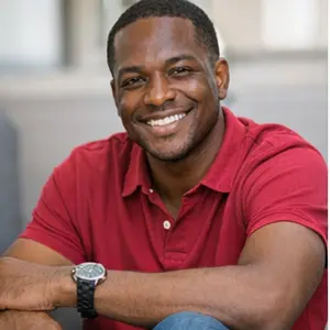 Ein lächelnder Mann mit rotem Polohemd und Armbanduhr sitzt mit auf die Knie gestützten Armen in einer entspannten, lässigen Pose - vielleicht beim Brainstorming für UG gründen.