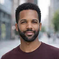 Ein Mann mit kurzen lockigen Haaren und einem gestutzten Bart lächelt sanft, während er im Freien auf einer Stadtstraße steht, vielleicht nachdem er Notardienstleistungen organisiert hat. Er trägt ein kastanienbraunes T-Shirt und der Hintergrund ist leicht unscharf.