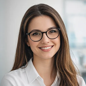 Eine Frau mit langen braunen Haaren und Brille lächelt in die Kamera. Sie trägt ein weißes Hemd und wartet möglicherweise auf ein Gespräch über die Sitzverlegung beim Notar in einem hell beleuchteten Innenraum mit unscharfem Hintergrund.