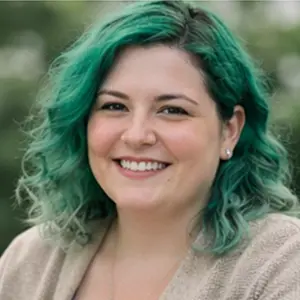 Eine Person mit gewelltem grünem Haar, die kleine Ohrstecker und einen beigen Pullover trägt, lächelt im Freien vor einem unscharfen grünen Hintergrund - vielleicht wartet sie auf Nachrichten über einen Geschäftsführerwechsel Notar Berlin.