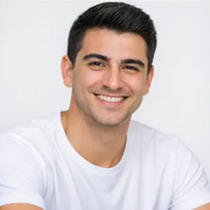 Ein junger Mann mit kurzen dunklen Haaren und hellem Teint lächelt in die Kamera. Er trägt ein schlichtes weißes T-Shirt, posiert vor einem einfachen hellen Hintergrund und repräsentiert Holding Gründen Berlins frisches neues Talent.