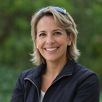 Eine Frau mit kurzen blonden Haaren, die eine dunkle Jacke und eine Sonnenbrille auf dem Kopf trägt, steht lächelnd im Freien vor einem unscharfen grünen Hintergrund und verkörpert die freundliche Professionalität eines Notars in Berlin.