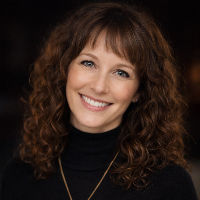 Eine Frau mit lockigem braunem Haar und Pony lächelt in die Kamera. Sie trägt einen schwarzen Rollkragenpullover und eine Halskette. Ihr Selbstbewusstsein strahlt - vielleicht zur Feier eines kürzlich erfolgten Grundschuldlöschens - vor einem dunklen, verschwommenen Hintergrund.